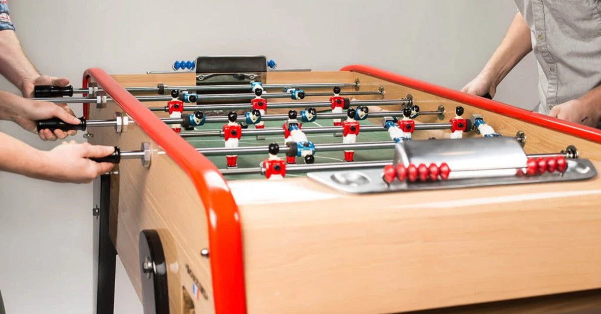 Three white people play table soccer or a light wood foosball table with red trimming along the edge.