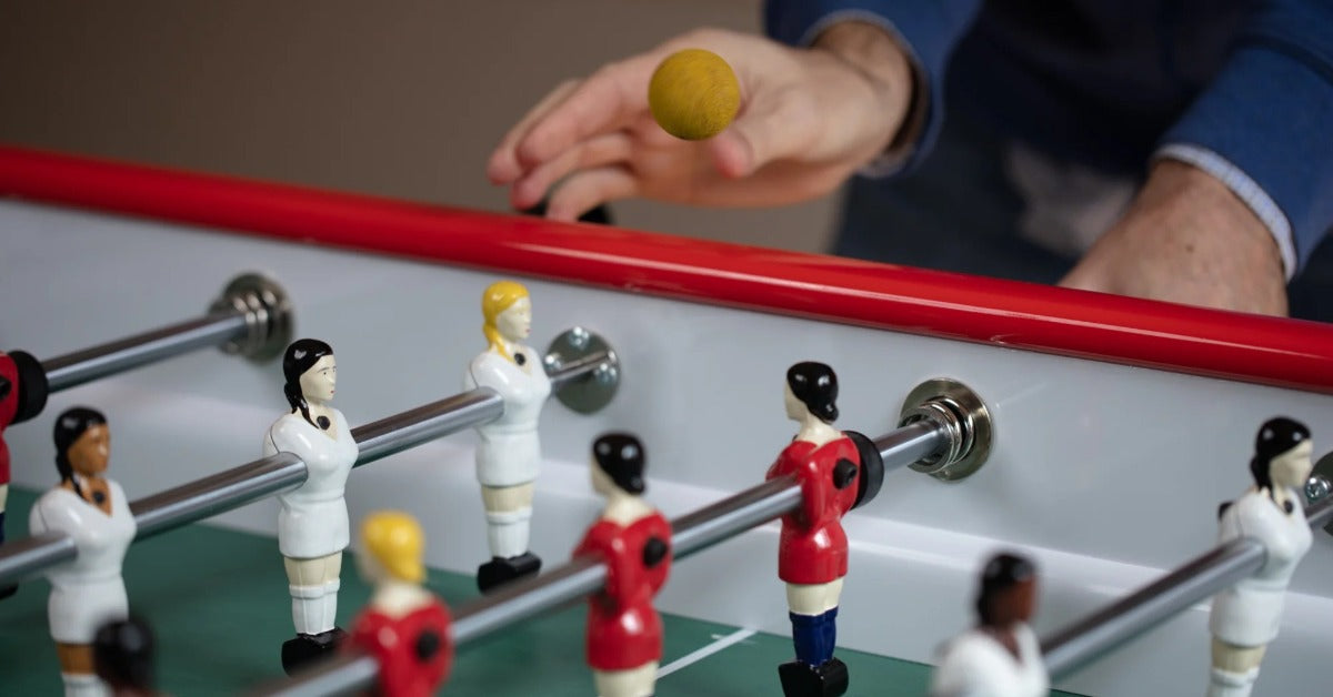A person's hand drops a foosball ball onto the table as players line up on rods during a casual indoor game.