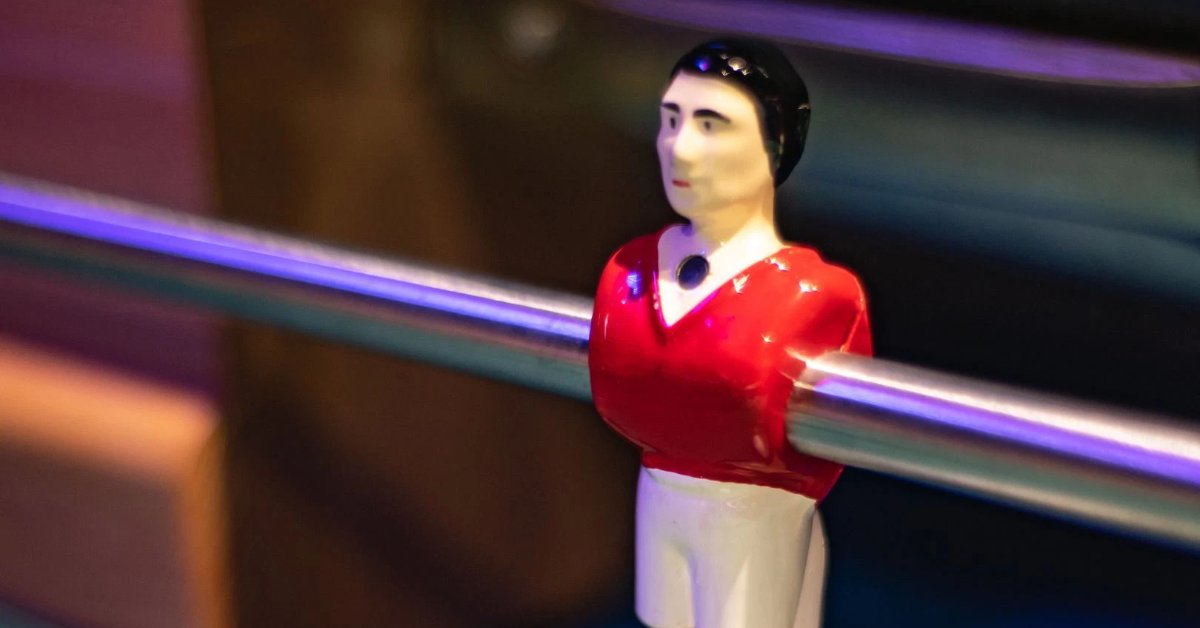 A close-up of a foosball player figure in a red jersey mounted on a metal rod, with a blurred game table background.